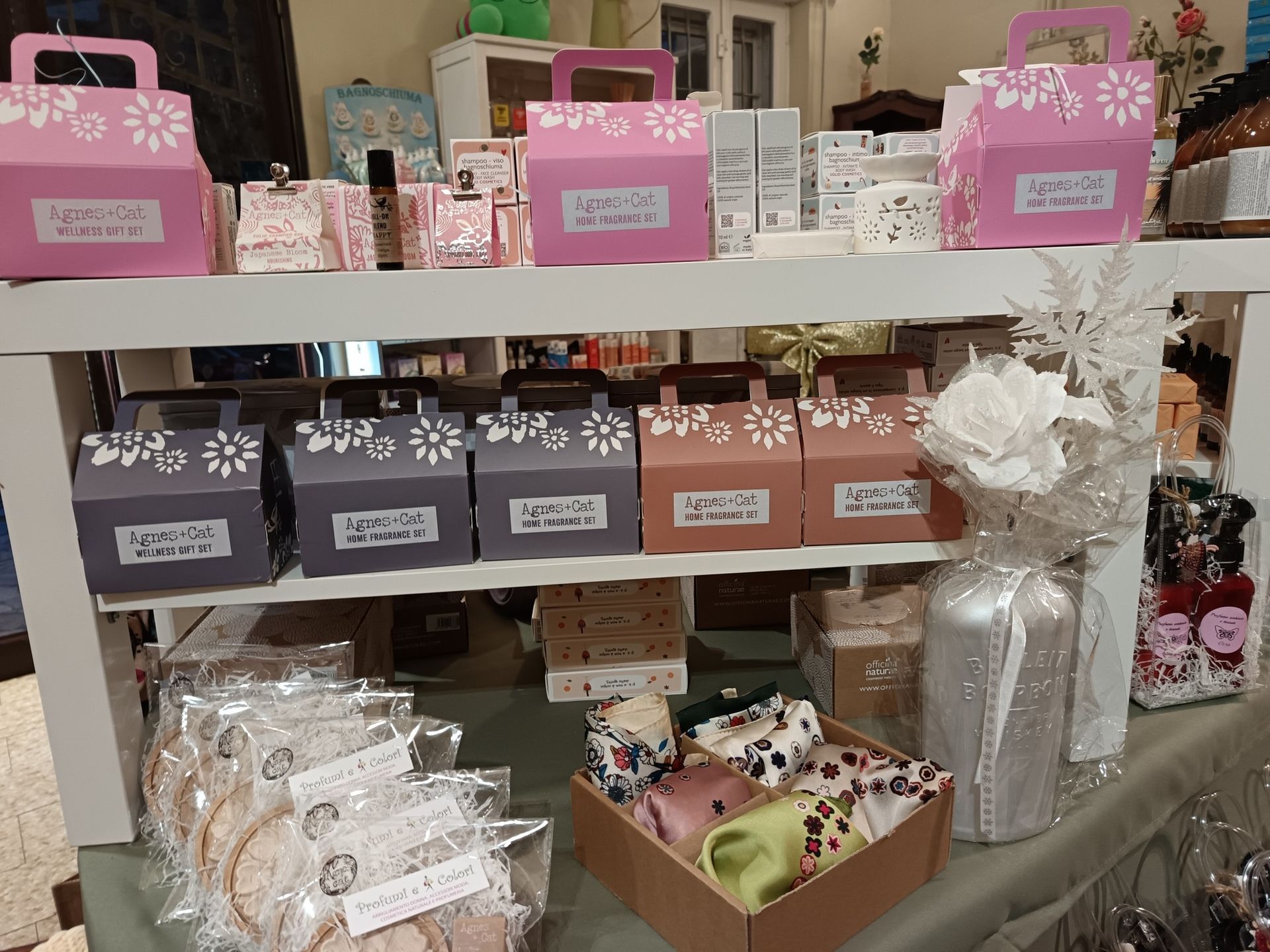 Scaffali con scatole regalo rosa, grigie e marroni. Alcune scatole hanno ritagli di fiori. Sono visibili anche biscotti e sciarpe.