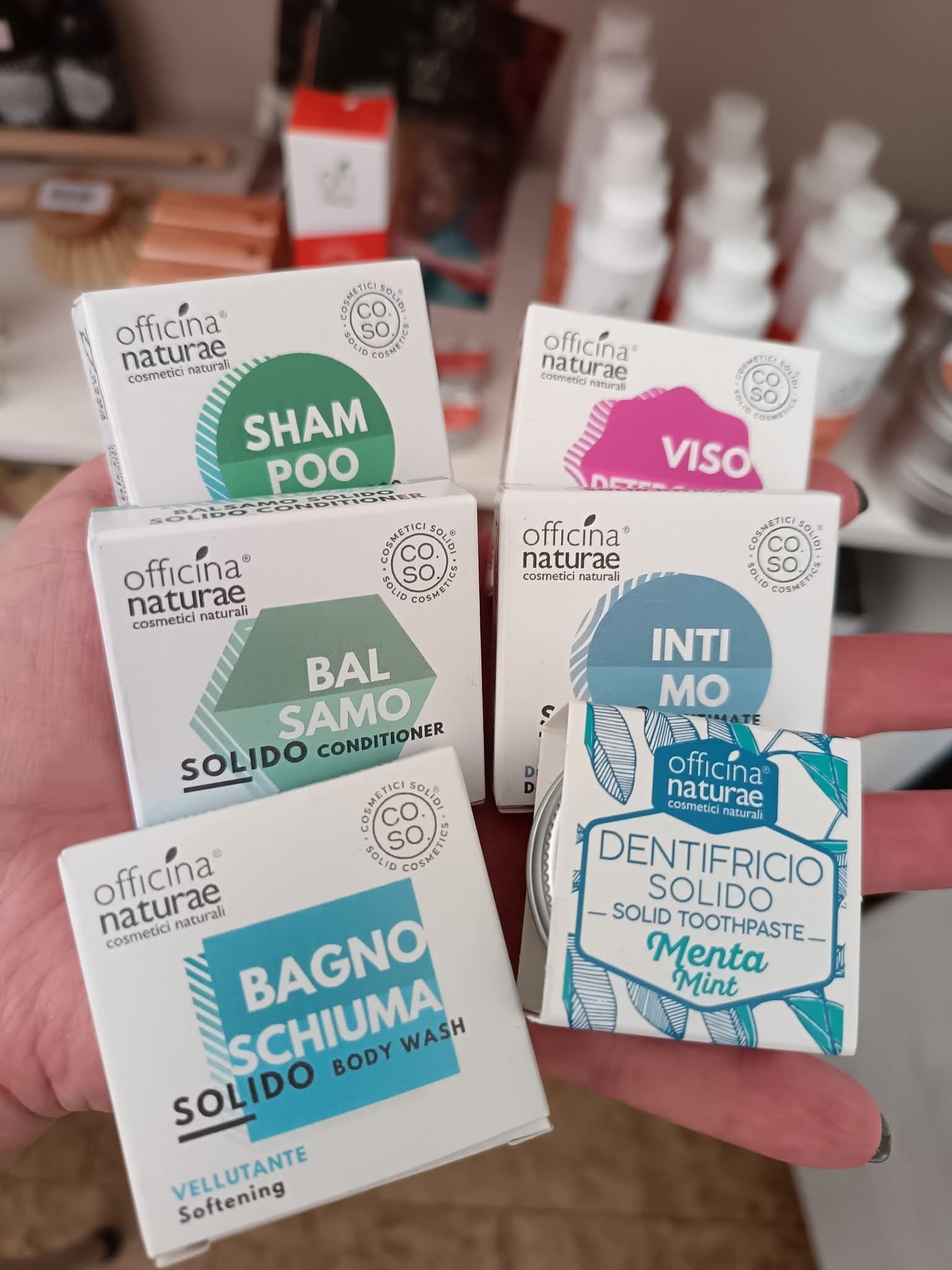 Mano che tiene in mano diversi piccoli prodotti da toeletta solidi ed ecologici, confezionati in una scatola: shampoo, detergente per il viso, bagnoschiuma, detergente intimo, dentifricio.