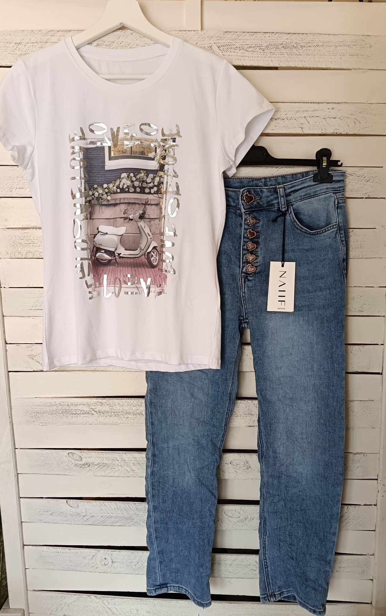 Maglietta bianca con motivo decorato, abbinata a jeans in denim chiaro con bottoni e sfondo in legno.