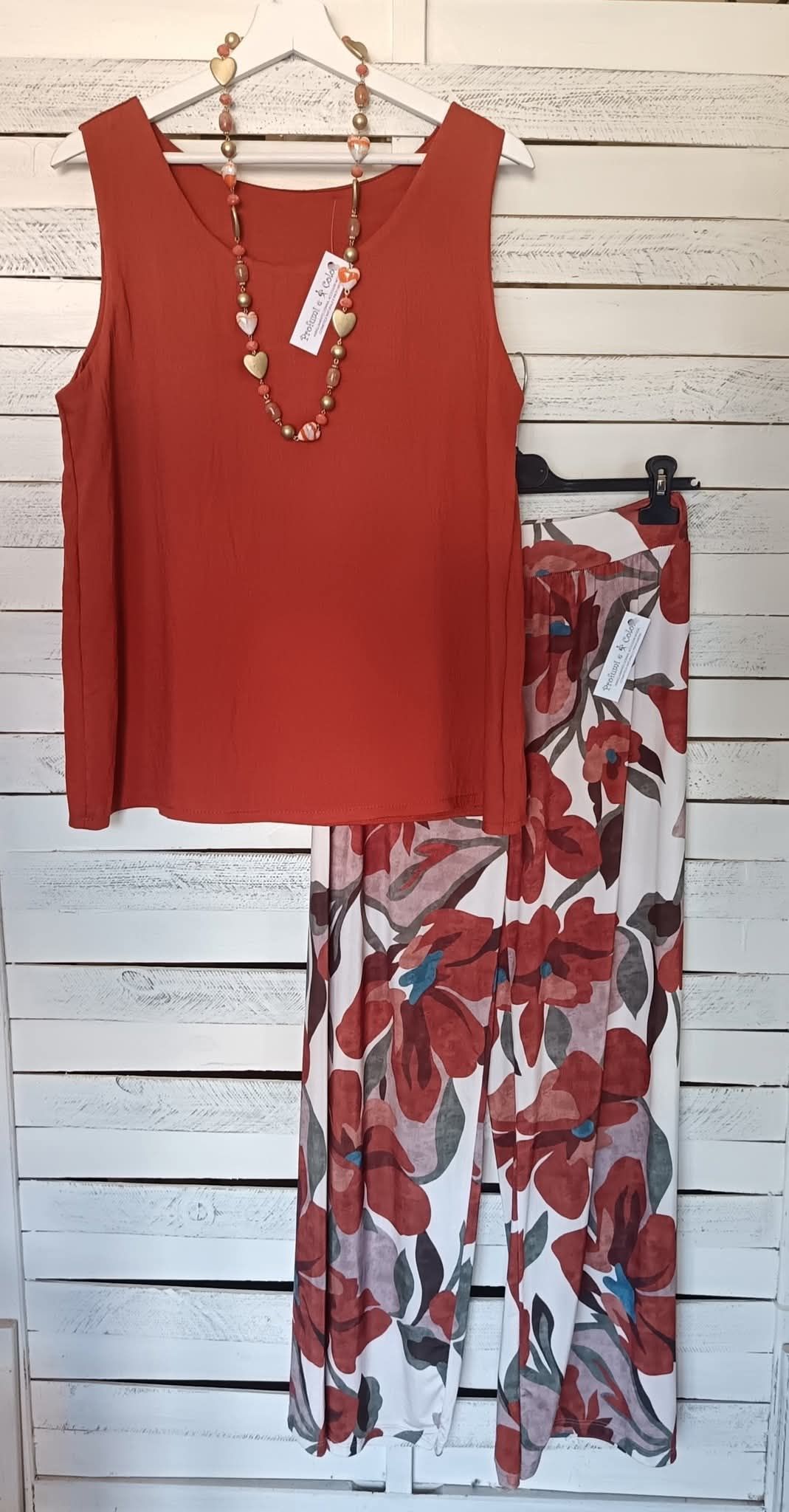 Completo composto da canotta color ruggine e pantaloni a fiori, appeso su un attaccapanni contro una parete imbiancata.