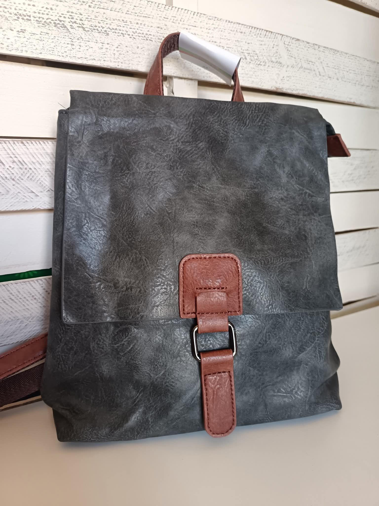 Zaino grigio con dettagli marroni, su uno scaffale bianco su uno sfondo di legno bianco.