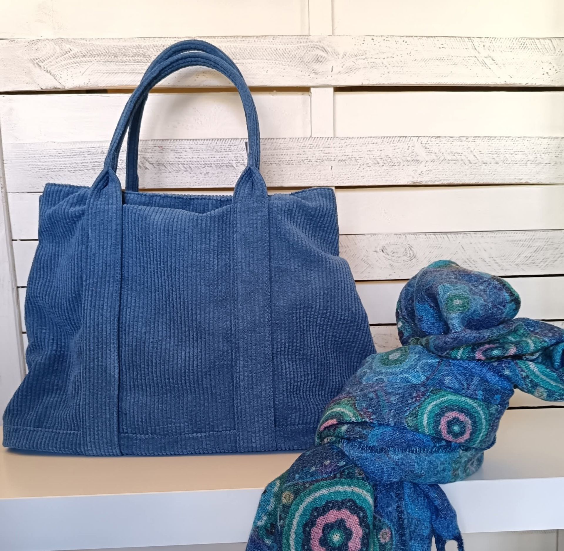 Borsa tote in velluto a coste blu e sciarpa fantasia su una superficie bianca contro uno sfondo in legno bianco.