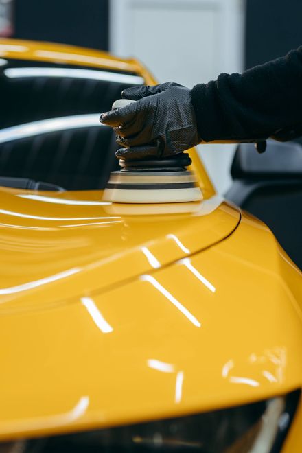 Person polishes a bright yellow car hood with an electric buffer, wearing black gloves.