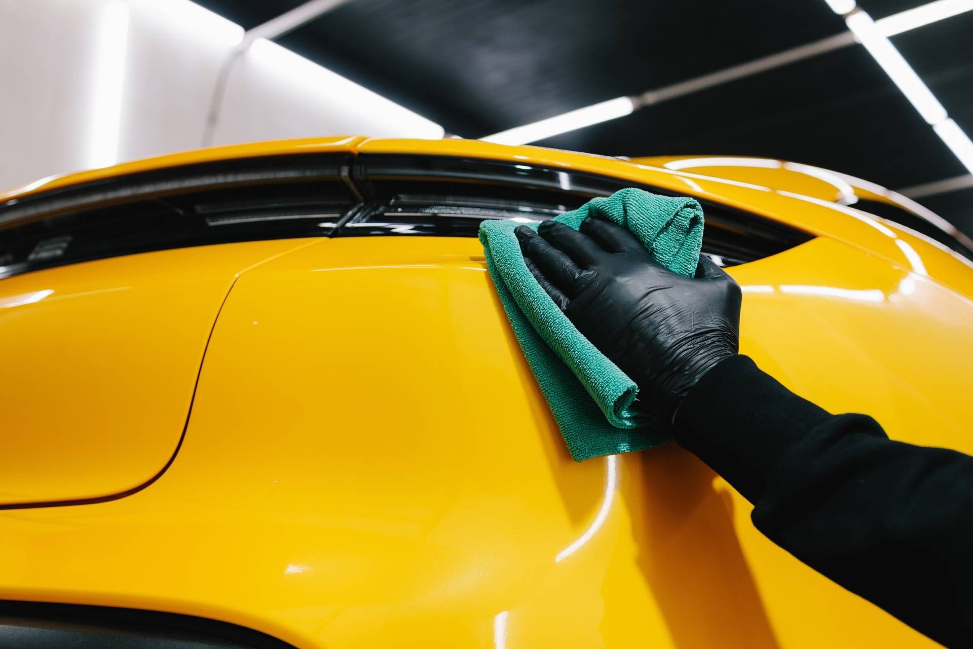 Person wearing black gloves wipes a yellow car with a green microfiber cloth.