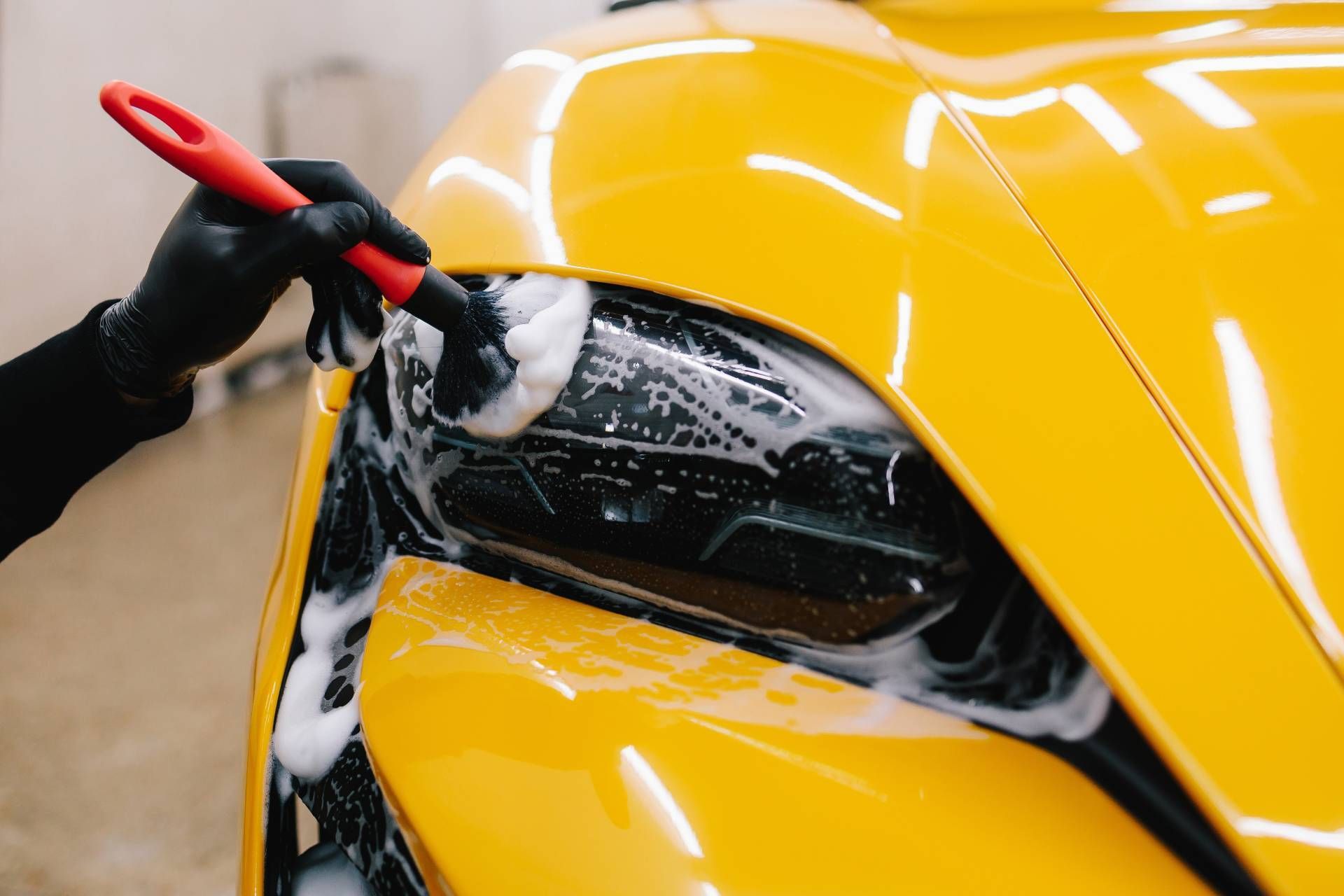 Person washing a yellow car headlight with soap and brush, wearing black gloves.