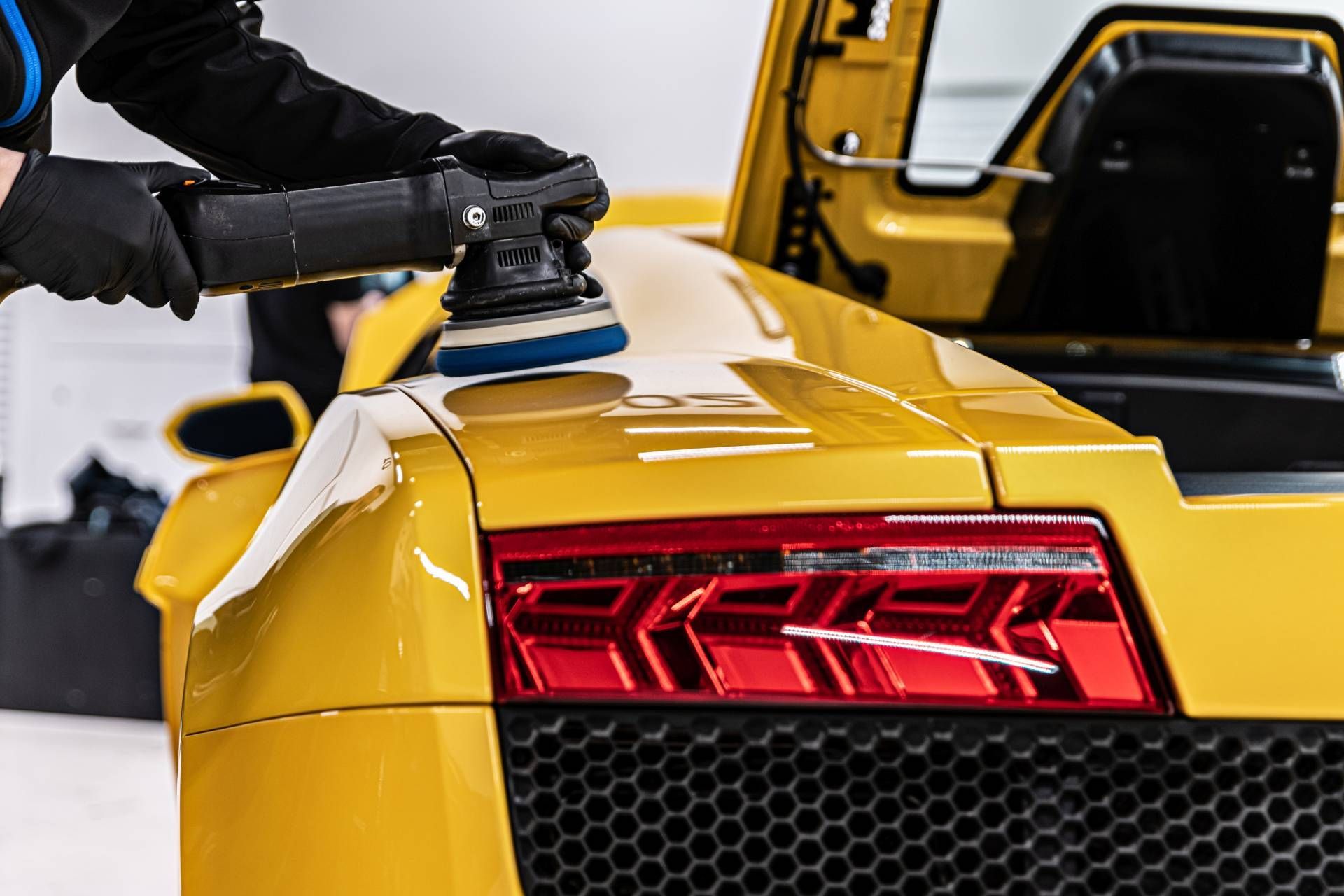 A person polishing a yellow sports car's rear with a machine polisher, with the trunk open.