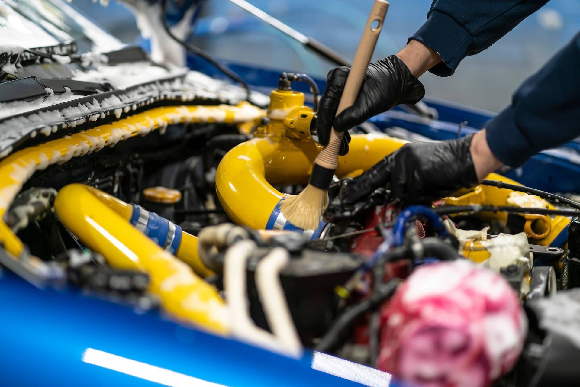 Person wearing black gloves cleaning a car engine with a brush; yellow and blue components.