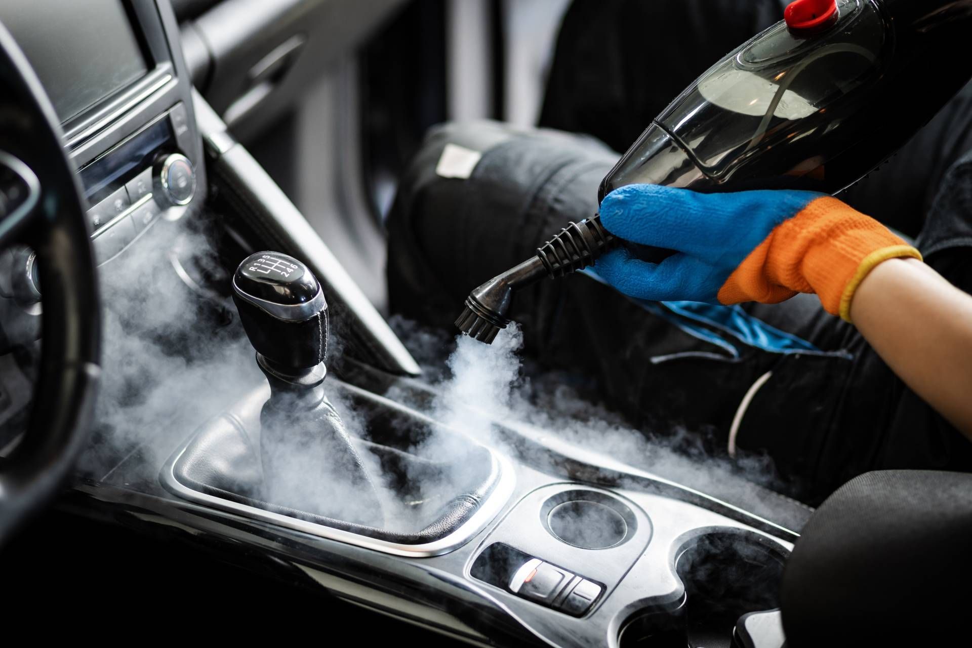 Person steam cleaning a car's interior using a steam cleaner, wearing gloves.