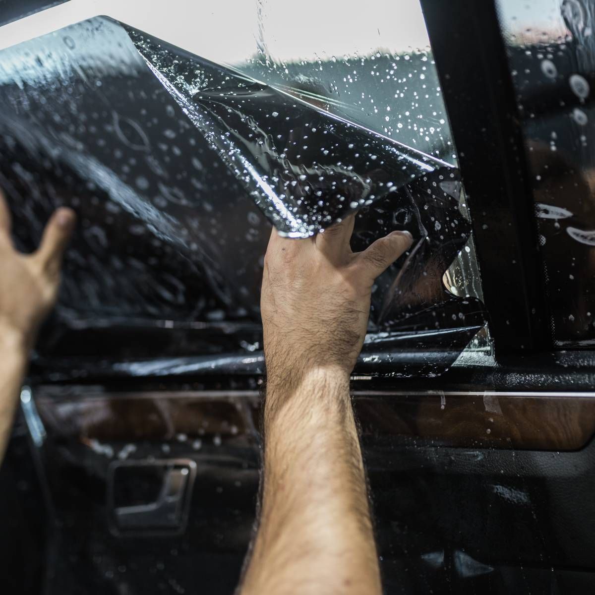 Hands applying tinted film to a car window. Film is dark, and the window frame is black.