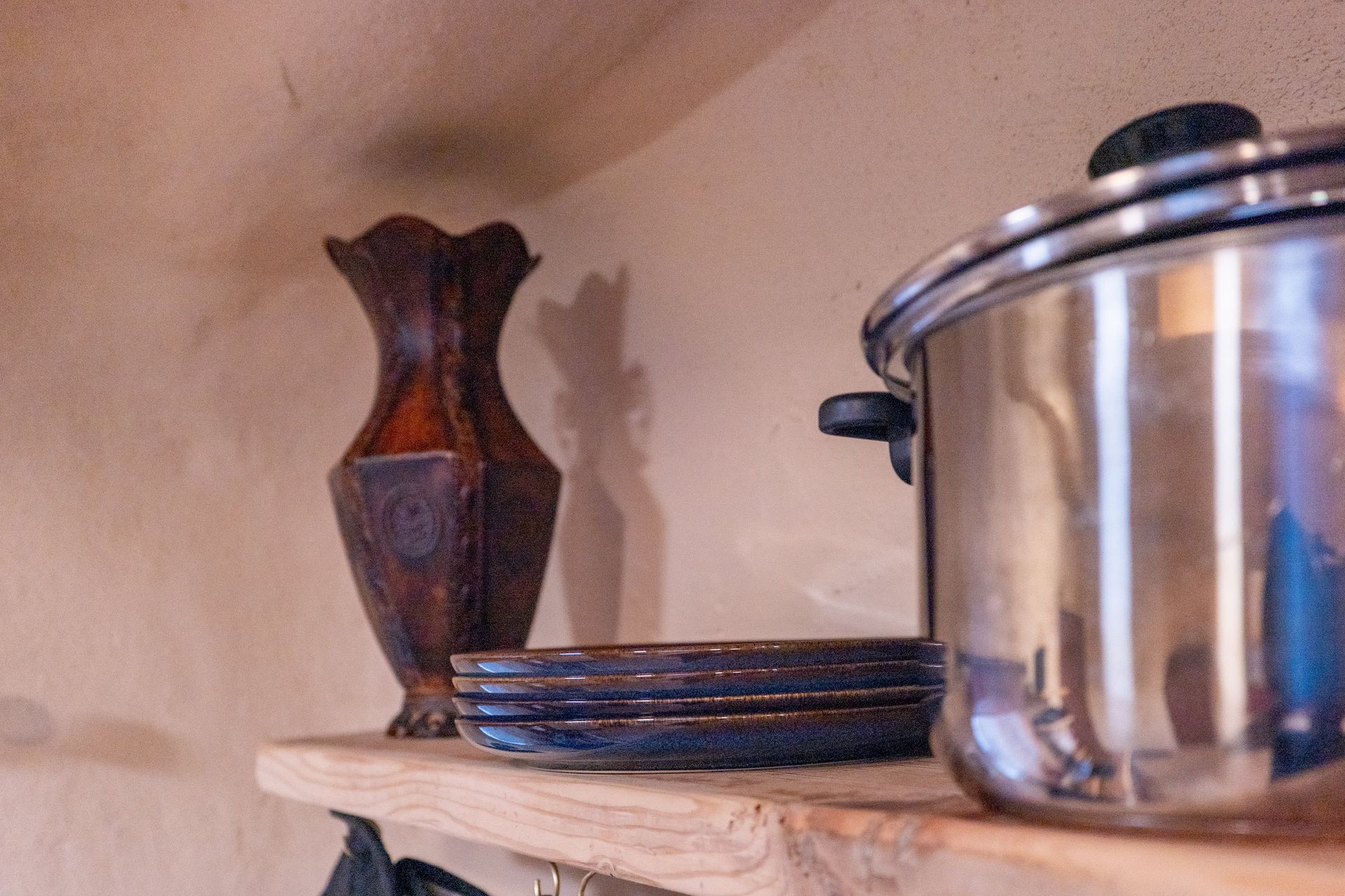 A pot and plates are on a wooden shelf next to a vase.