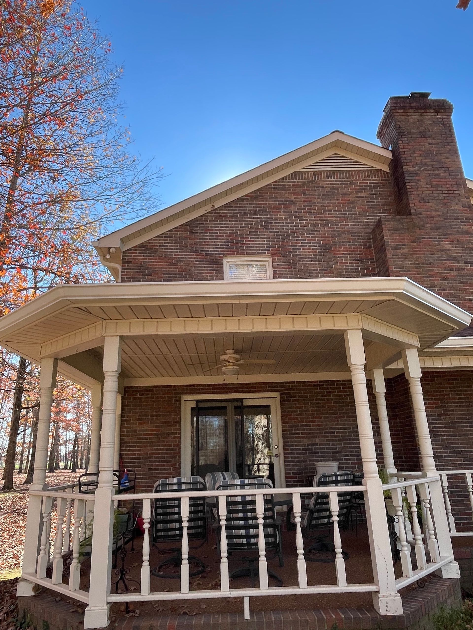 Brick house with a porch; trees with fall foliage in the background; clear blue sky.