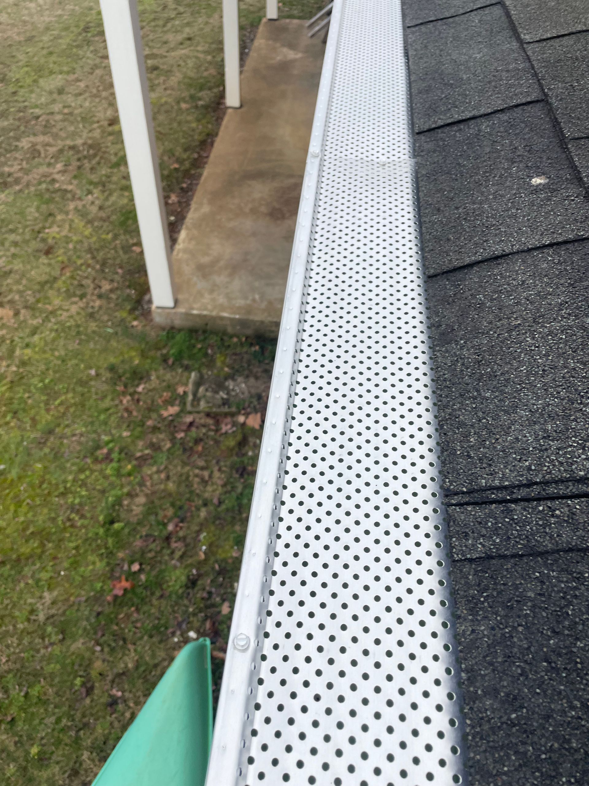 Gutter covered with a perforated metal guard next to a shingled roof and a concrete porch.