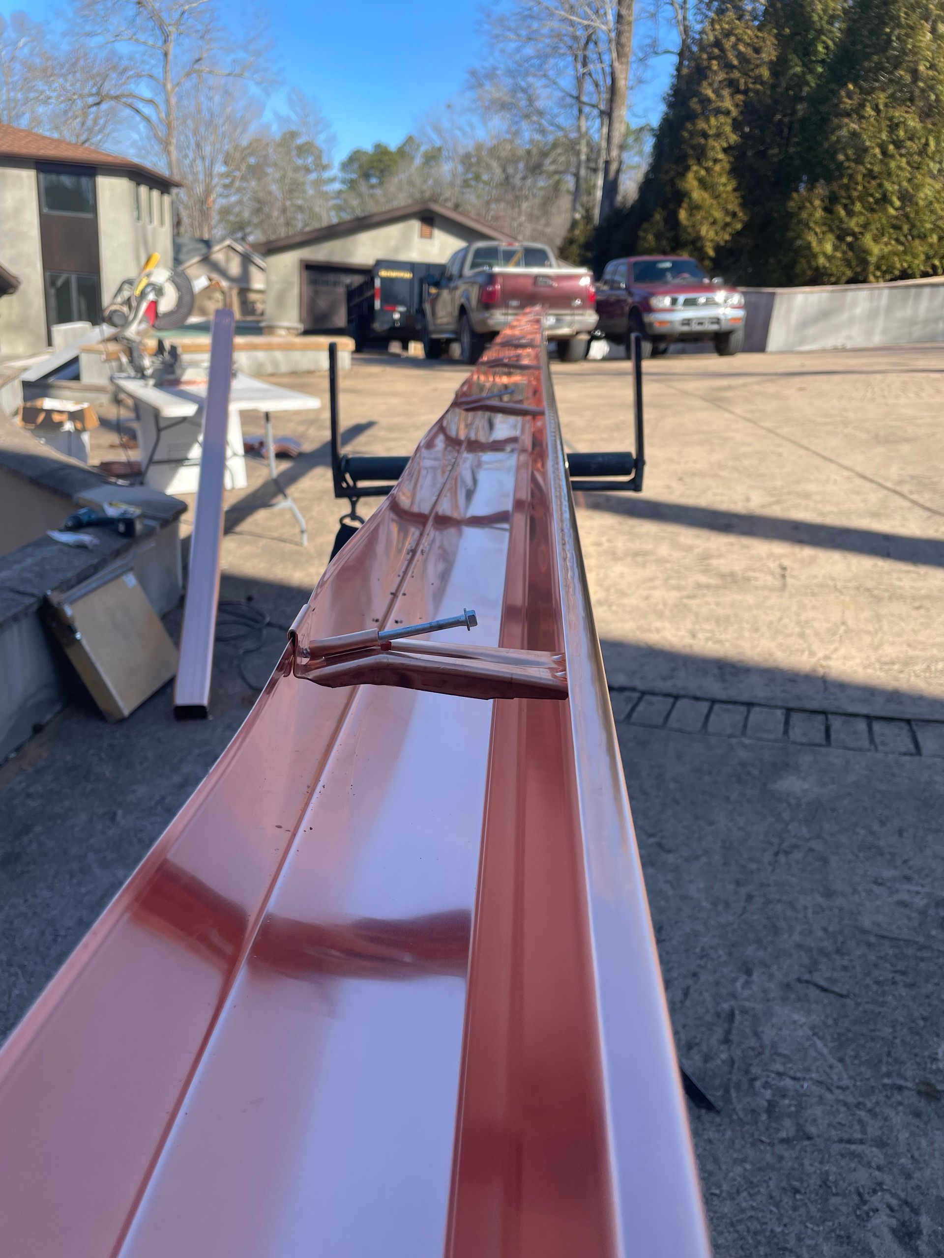 Long, copper-colored gutter segment on a black support, with a saw and vehicles in the background.