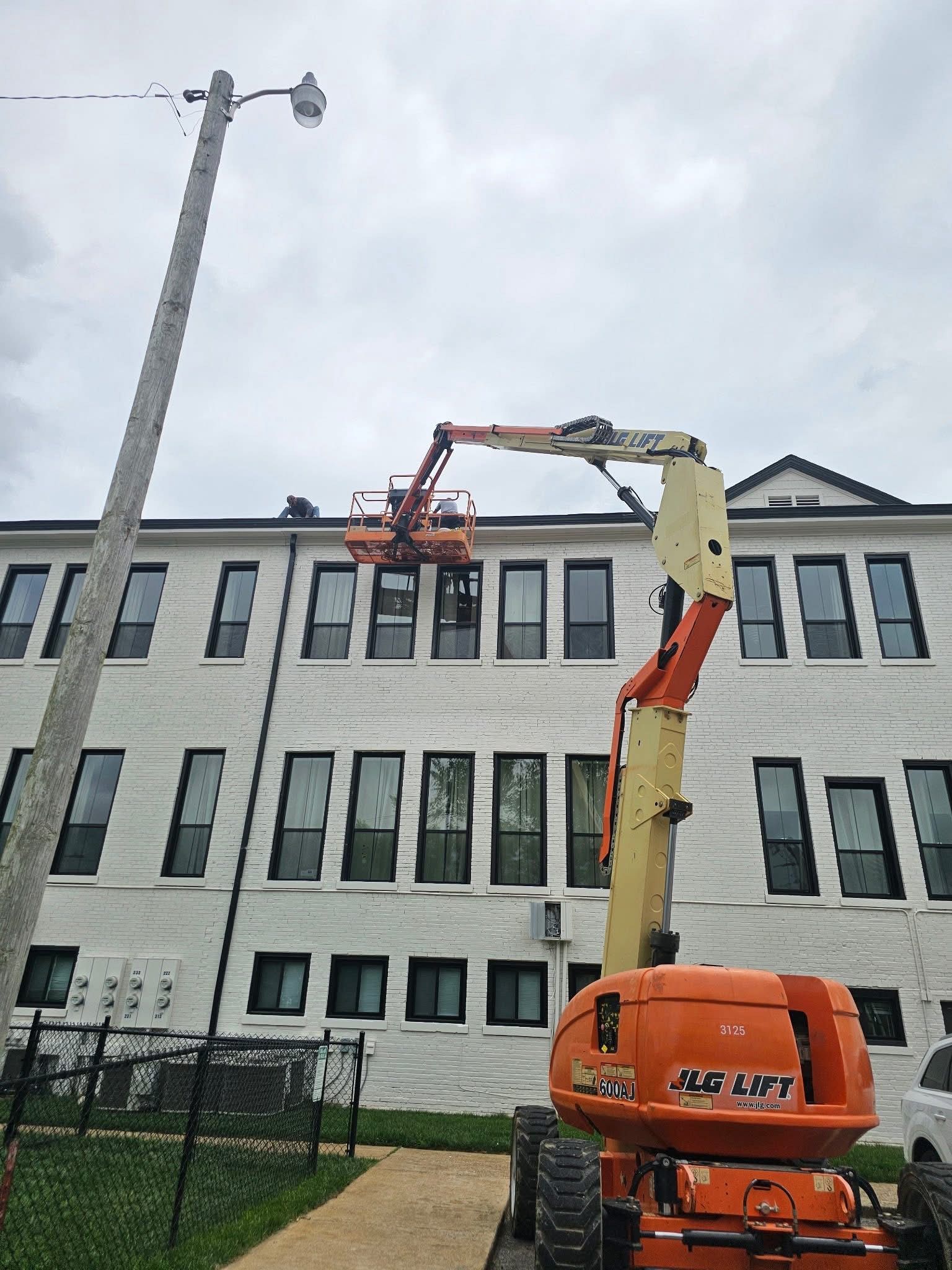 Orange lift with person near white building working on a window; gray sky.
