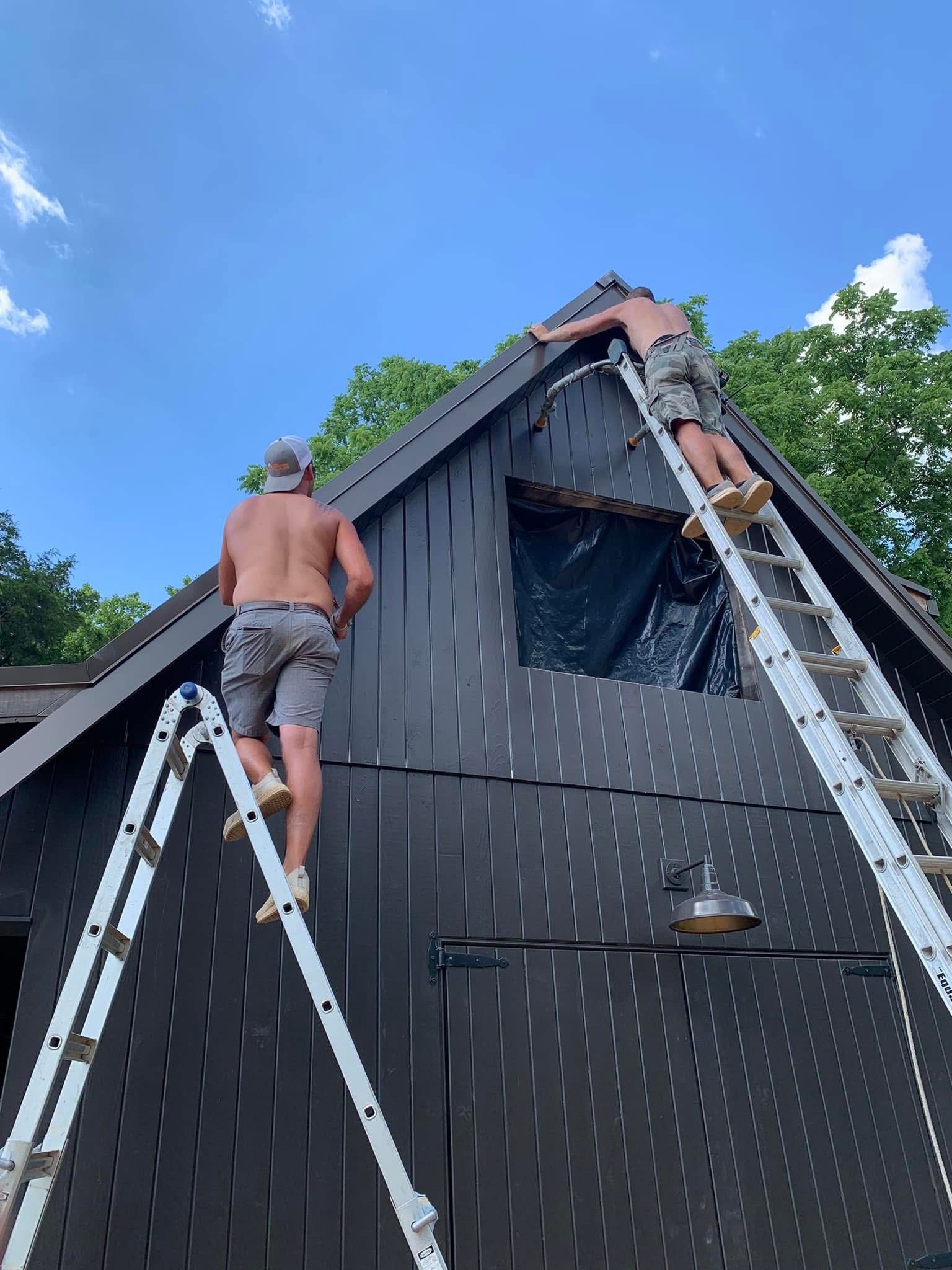 Two men on ladders working on a dark-painted A-frame building under a blue sky.