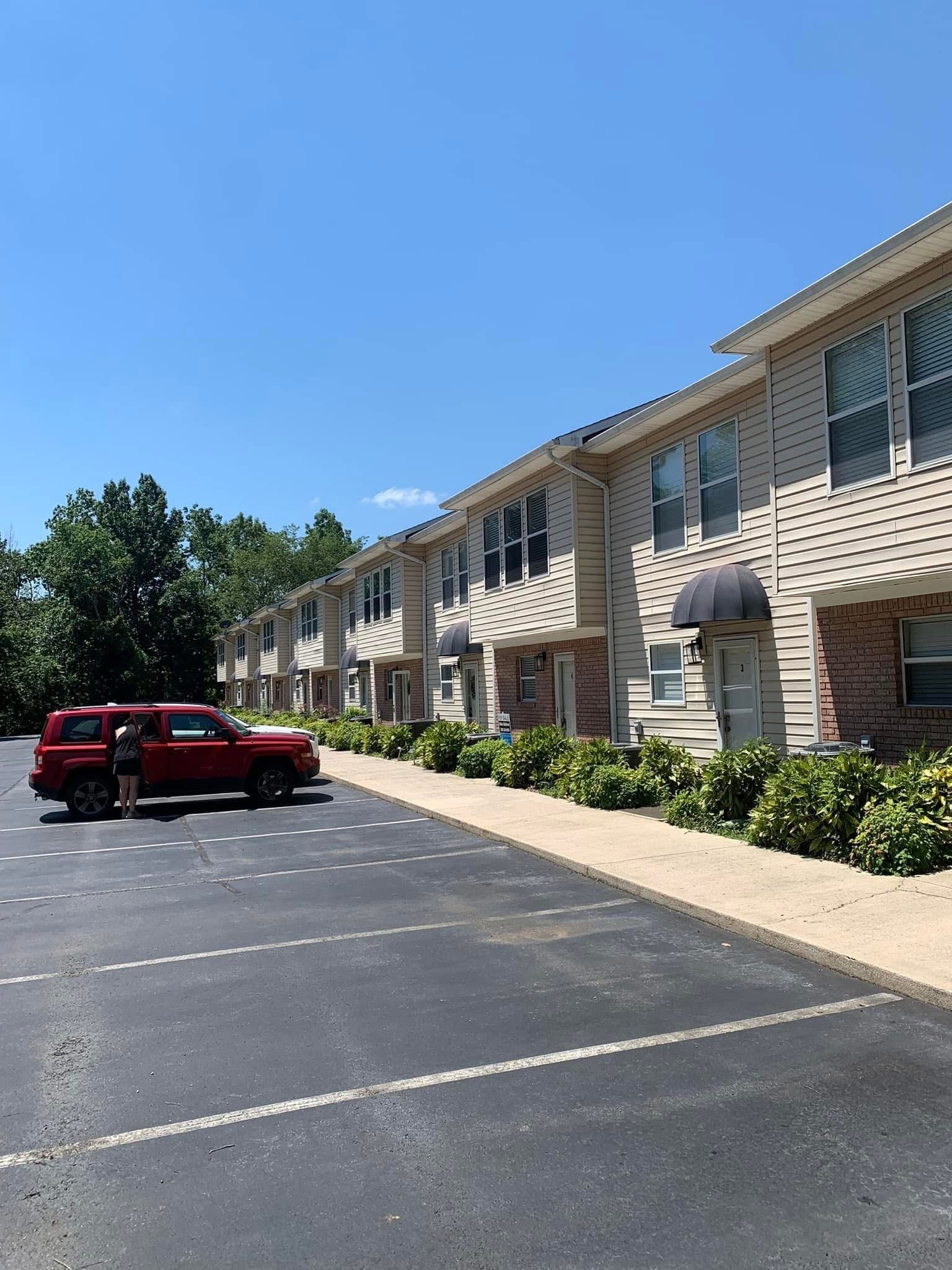 Red SUV parked in front of a row of two-story beige apartment buildings with a blue sky overhead.
