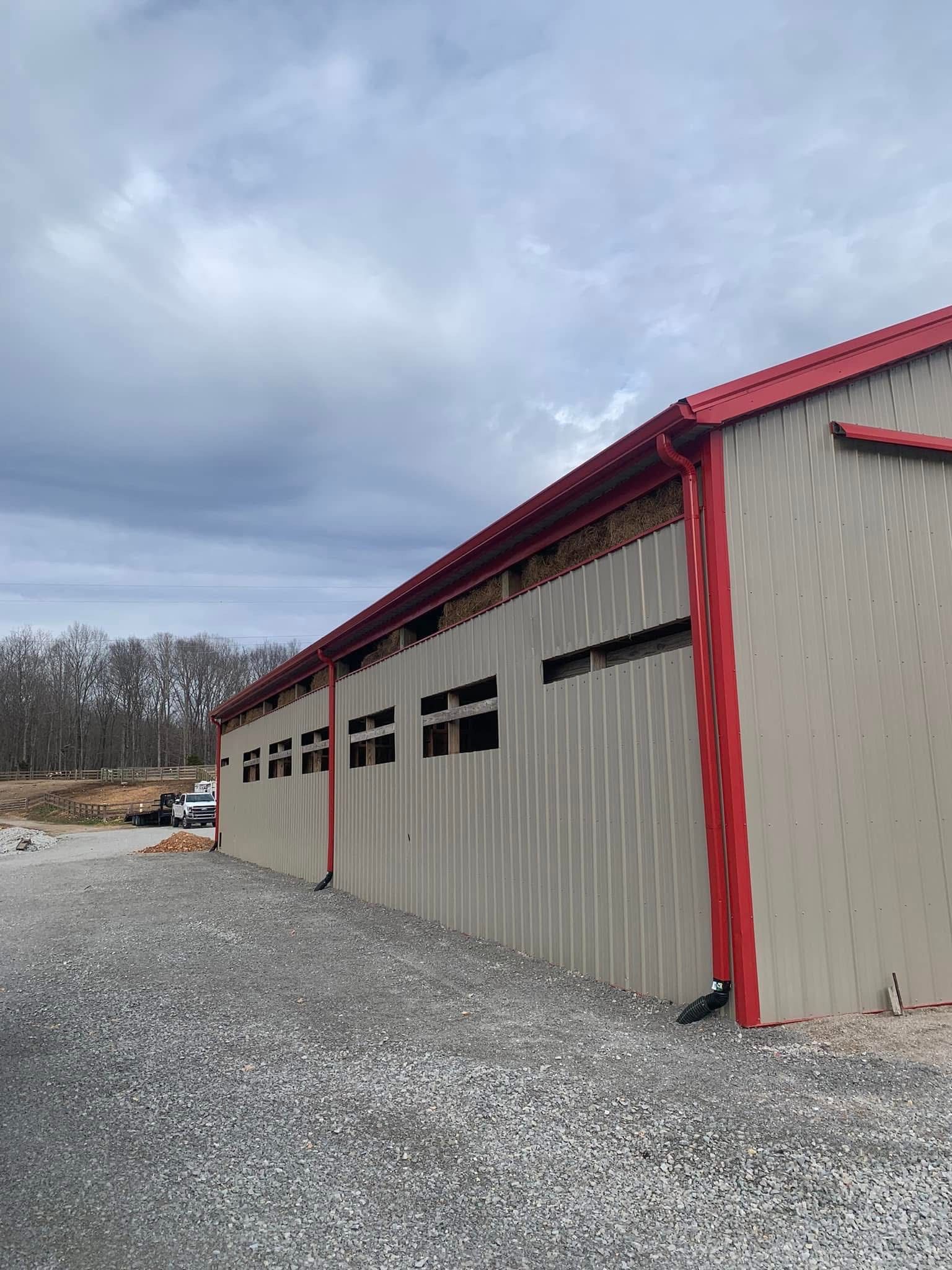Gray metal building with red trim and roof against cloudy sky. Gravel ground.