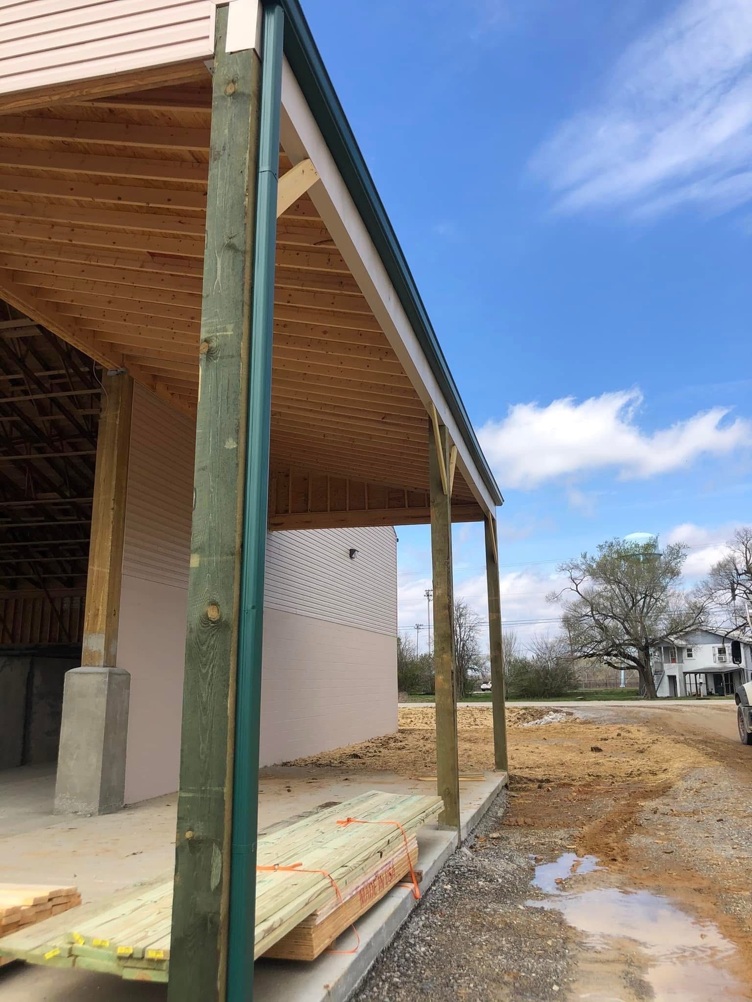 Construction site with a covered porch and lumber stacked. Green-wrapped posts, blue sky, and a dirt road.