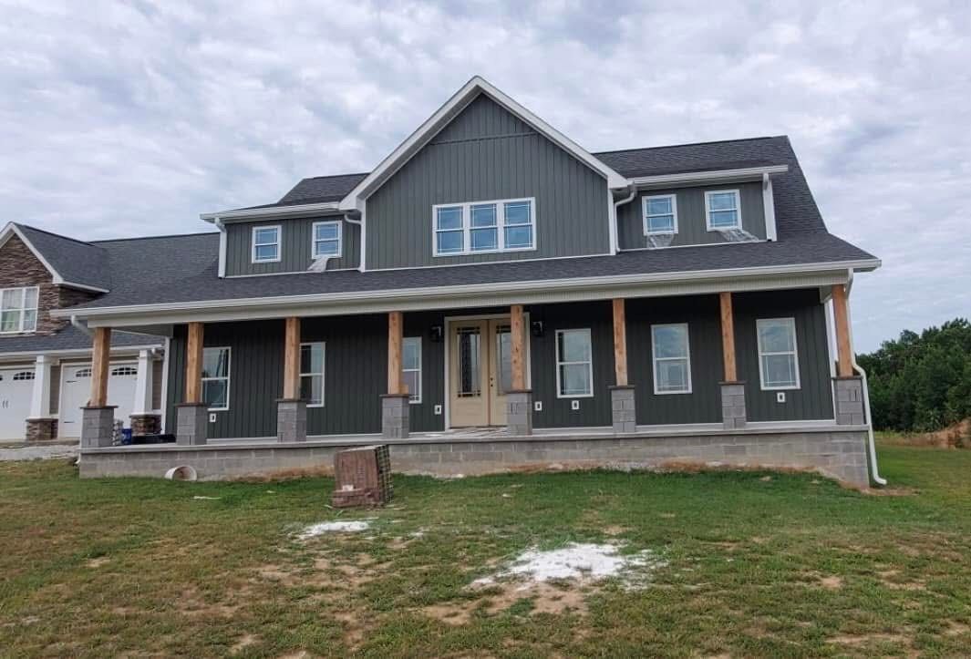 Two-story gray house with wrap-around porch, natural wood columns, and green lawn under cloudy sky.