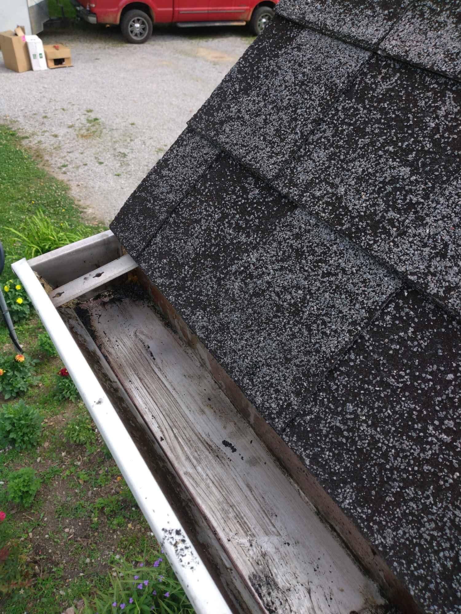 Gutter filled with debris next to a roof with black asphalt shingles; red truck in background.