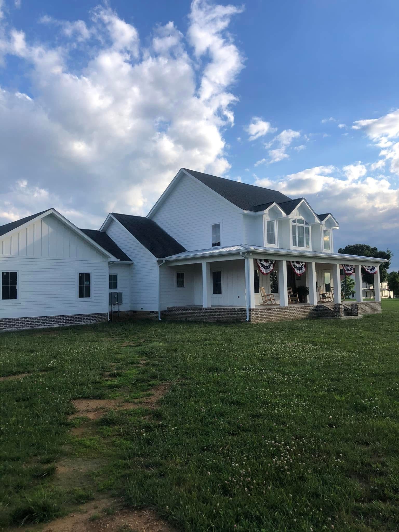 White farmhouse with a porch, black roof, on a green lawn under a cloudy blue sky.