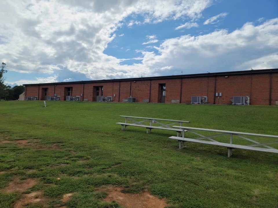 A long brick building with multiple doors, grass, and bleachers under a cloudy sky.