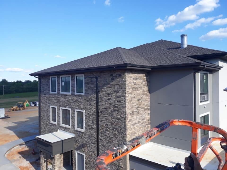 Two-story building under construction with gray stone facade, gray siding, and dark roof, viewed from an elevated work platform.