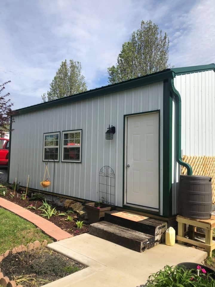 White metal shed with green trim, door, and small windows, steps to the door, rain barrel.