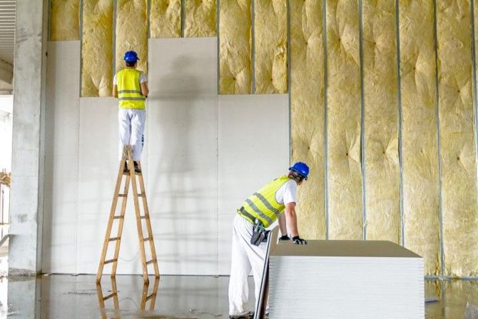 Two workers in hard hats install insulation in an unfinished interior wall.
