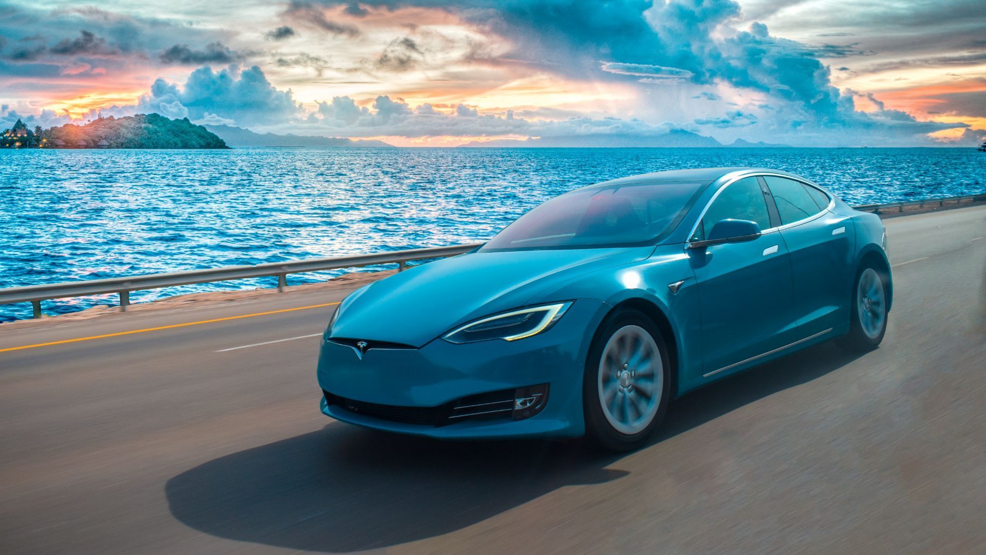 a blue Tesla model S  electric vehicle is driving down a road next to the ocean.