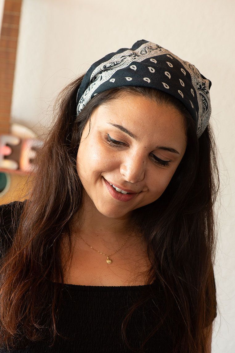 Frau mit Kopftuch, lächelnd, den Blick gesenkt; in der Nähe einer hellen Wand, im Hintergrund ist eine Gitarre zu sehen.