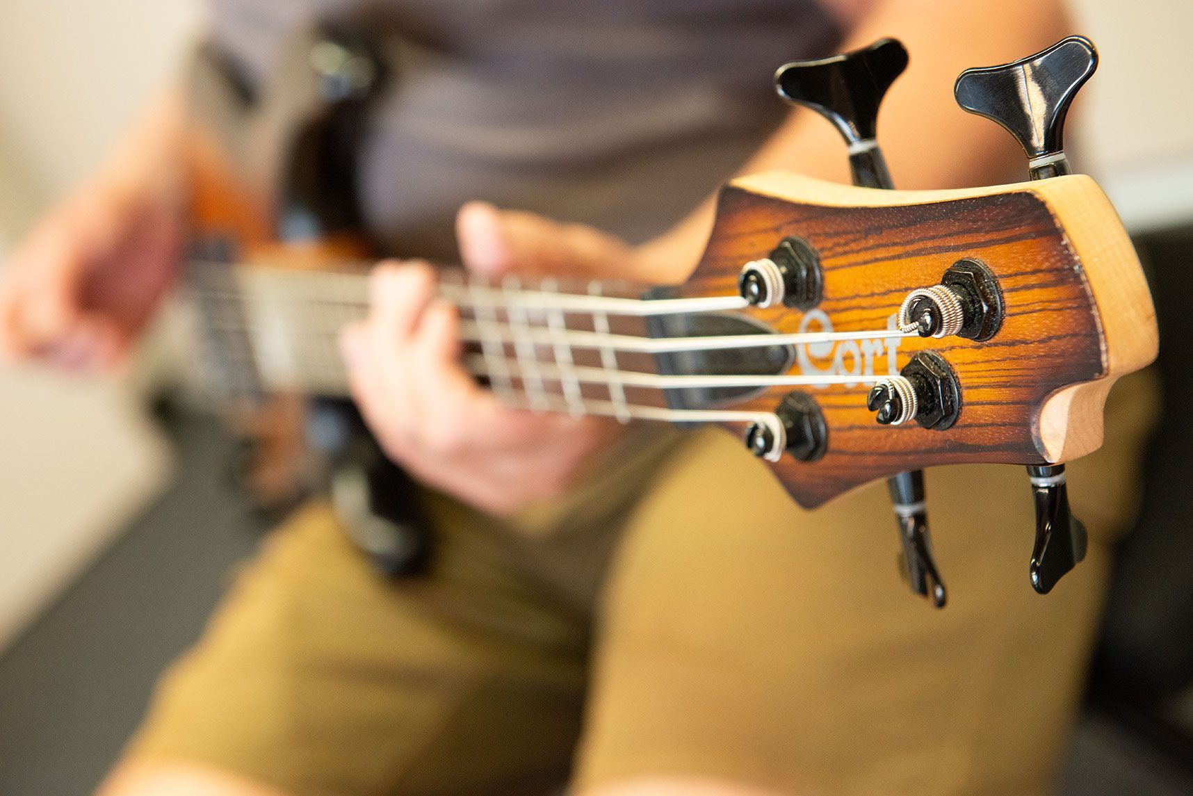 Nahaufnahme einer Bassgitarrenkopfplatte mit Stimmwirbeln; im Hintergrund spielt eine Person das Instrument.