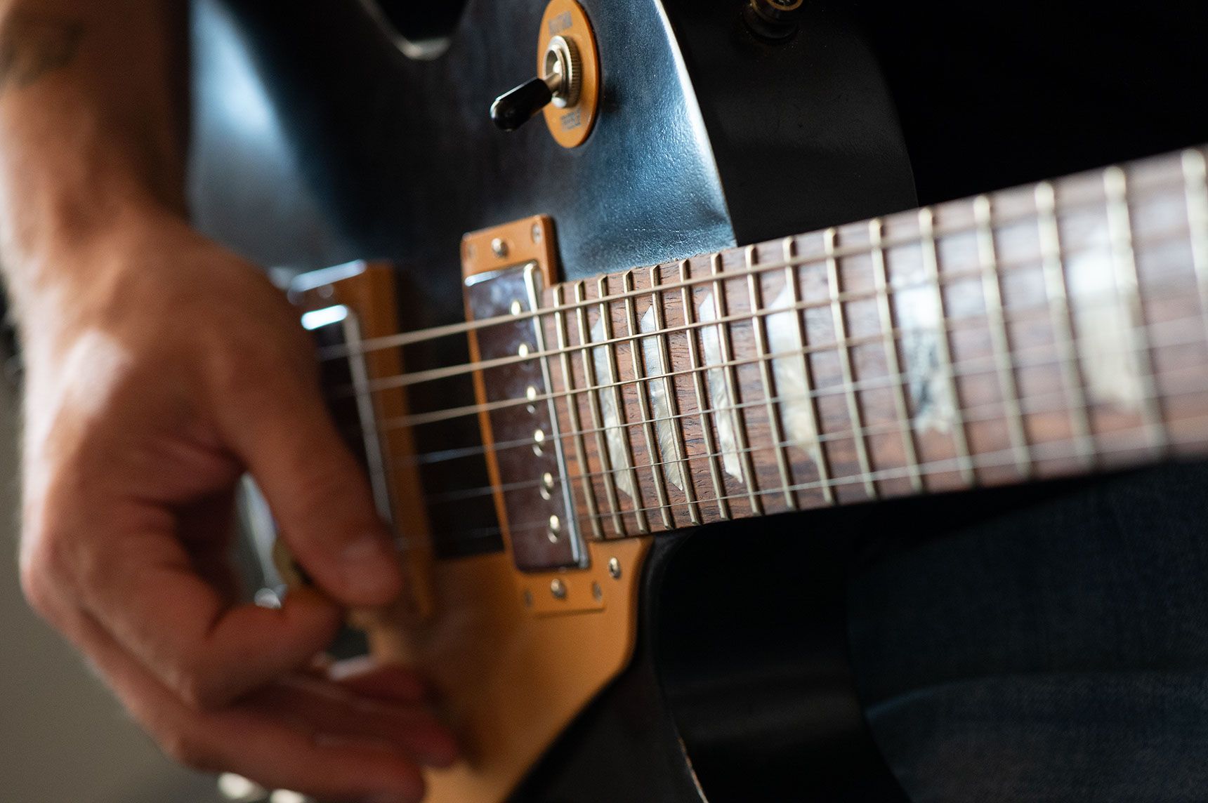 Nahaufnahme einer Hand, die eine schwarze E-Gitarre anschlägt; Bünde sichtbar, goldene Hardware.