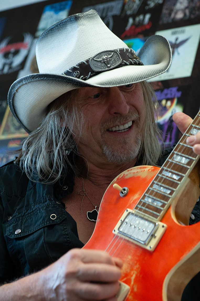 Mann mit langen Haaren und Cowboyhut, der eine orangefarbene E-Gitarre hält, lächelt.