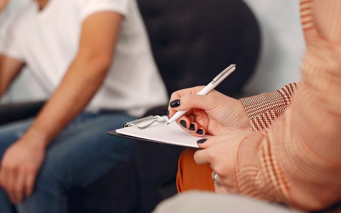 A woman is writing on a clipboard while a man sits in the background.