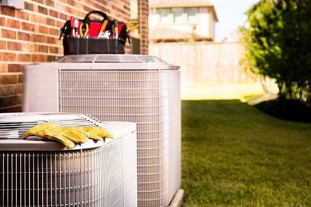 Two air conditioning units with a toolbox and gloves on top, in a grassy backyard.