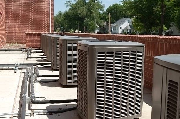 Row of air conditioning units on a rooftop next to a brick wall. Blue sky and trees in background.