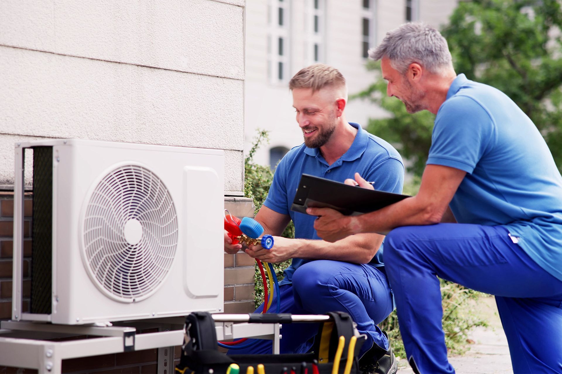 Two HVAC technicians examine an outdoor unit; one holds gauges, other looks at clipboard.