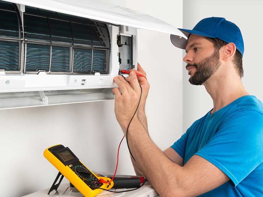 HVAC technician uses multimeter on an air conditioner. Blue shirt, baseball cap, and white room.