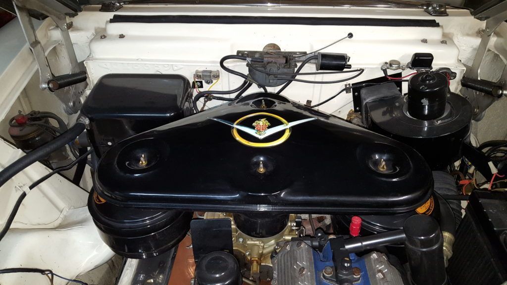 Engine compartment of a vintage white car with a black air cleaner featuring a gold emblem, and gold-colored engine components.