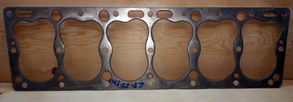 A metal head gasket with six large, irregular-shaped holes, likely for a vehicle engine.