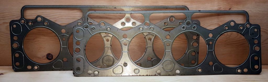 Two used engine head gaskets, possibly for a four-cylinder engine, on a wooden surface. They are metal, showing wear.