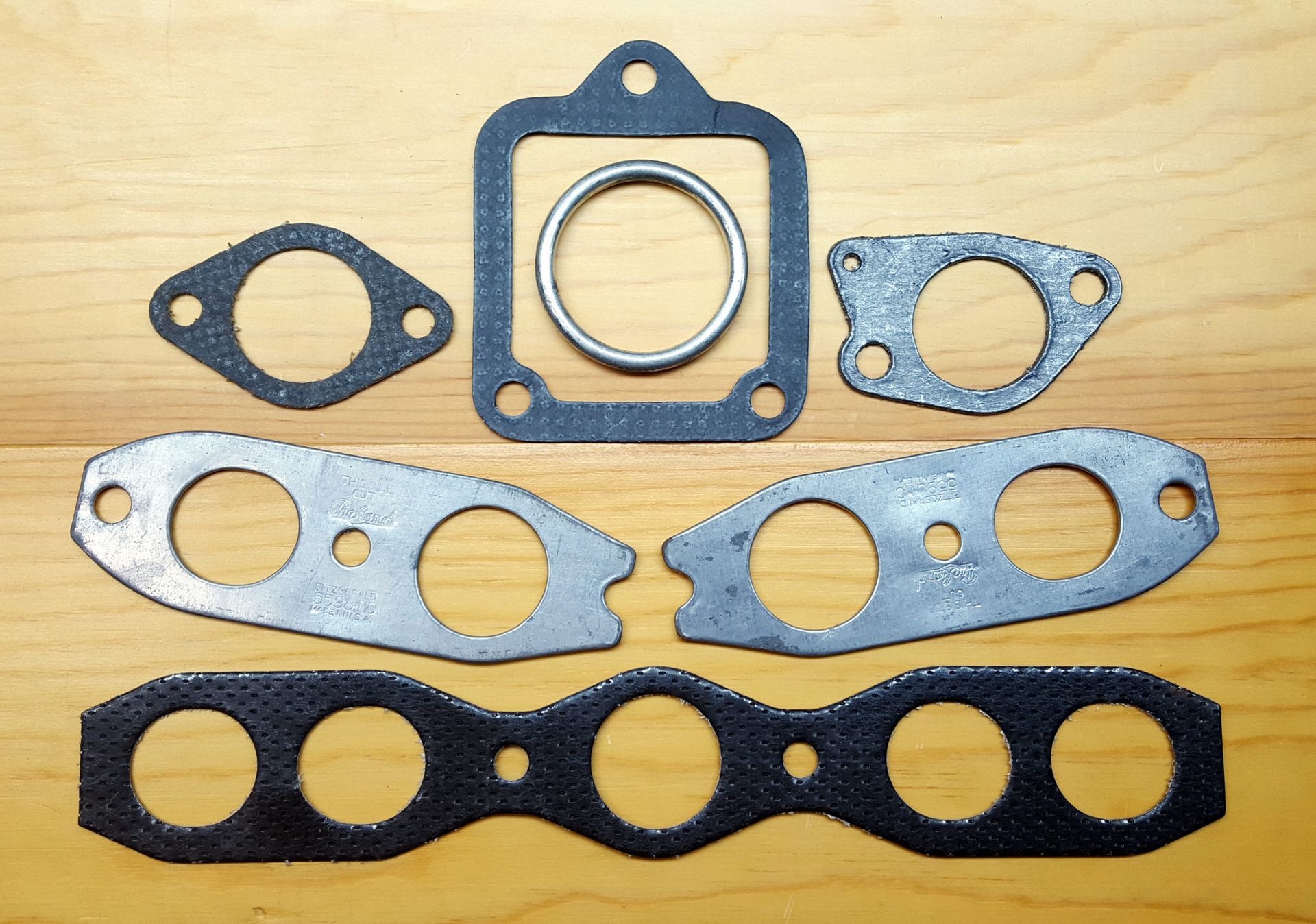 Various automotive exhaust gaskets and rings laid out on a wooden surface. Different shapes and sizes, mainly black and silver.