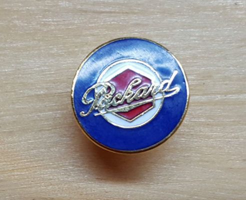 A blue, white, and red Packard logo pin with gold trim, set against a light brown wood background.