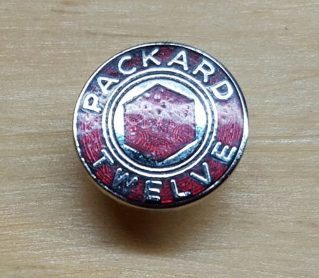 A vintage Packard Twelve emblem. The round, silver badge has red enamel and reads 