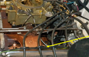 Close-up of a car engine with a carburetor, spark plug wires, and a distributor. Yellow arrow points to spark plug wire holder.