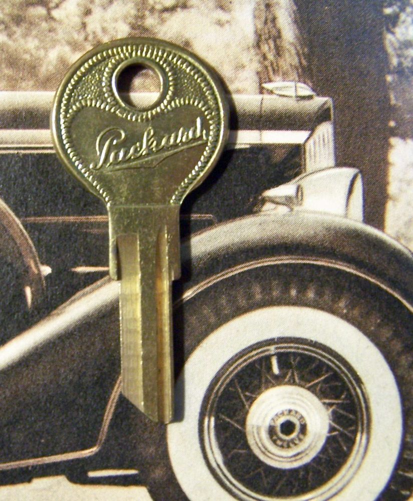 A brass Packard key with the logo, in front of a vintage car tire and fender.