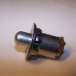 Silver push-button lamp with a domed end, surrounded by a mounting flange and electrical contacts.