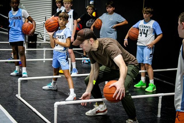 Coach demonstrates basketball dribbling drill; boys watch on court.