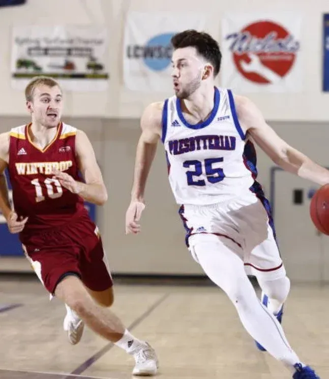 Basketball players in action. A Presbyterian player dribbles, pursued by a Winthrop player.